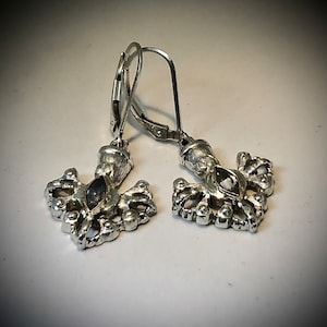 May include: A pair of silver-toned earrings. Each earring features a decorative, openwork design with a central, dark-colored stone. The earrings have lever-back closures. The design is ornate and detailed, with a vintage or antique aesthetic.