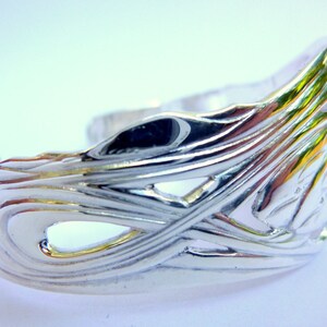 May include: A silver bracelet with a detailed, intricate design of overlapping waves. The bracelet is open and has a smooth, polished finish.