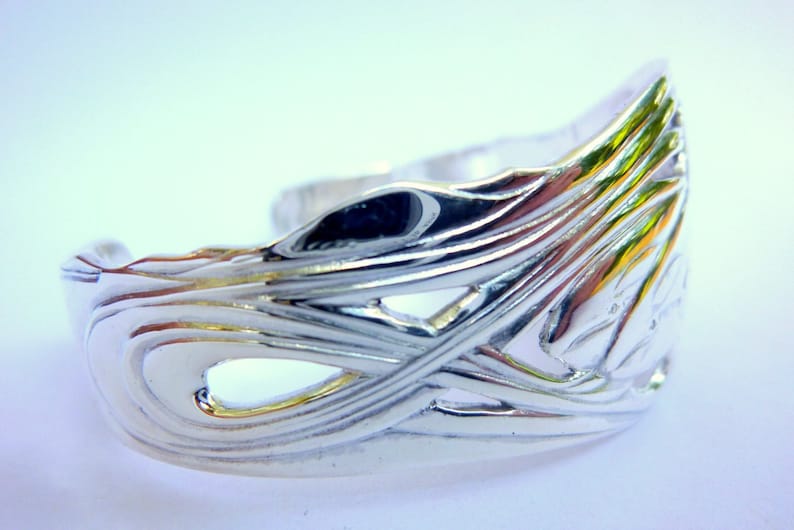 May include: A silver bracelet with a detailed, intricate design of overlapping waves. The bracelet is open and has a smooth, polished finish.