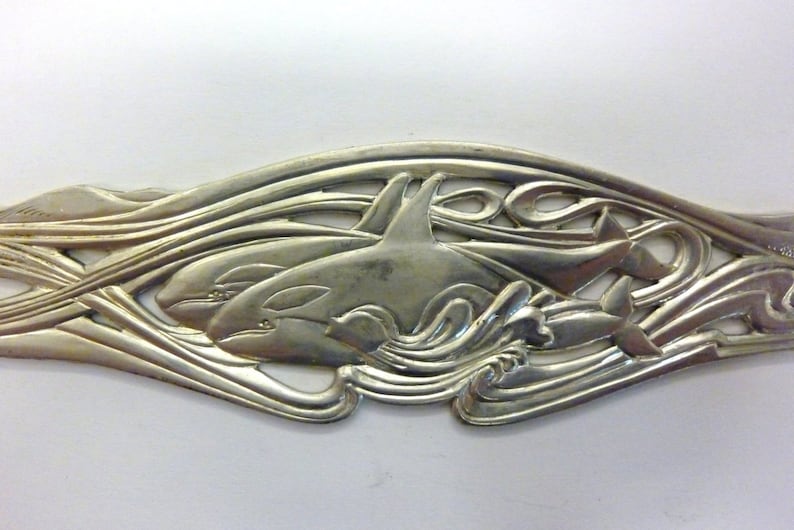 May include: Silver-toned metal ornament featuring two orcas swimming through stylized waves. The orcas are detailed with realistic features and the waves are intricate and flowing.