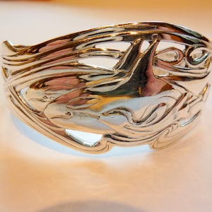May include: Silver bracelet with a detailed design of two dolphins swimming in waves.