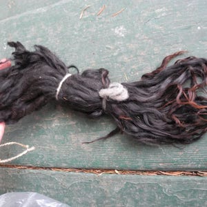 May include: A bundle of dark, wavy hair tied with white string and a gray piece of fiber. The hair is primarily black with some reddish-brown highlights. The hair is held by a person's hand, resting on a green wooden surface.