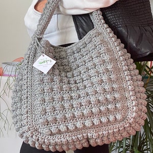 Op de afbeelding: Een grijze gehaakte tote bag met een textuur, een bolletjes design. De tas heeft een enkele band en een klein wit label met de tekst "Shopfully Handmade Bags".