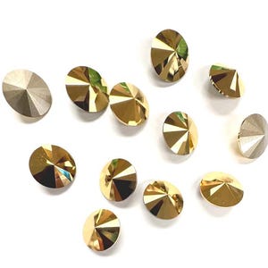 May include: An assortment of faceted, round-shaped crystal rhinestones in shades of gold and silver. The crystals have a multi-faceted design, reflecting light. The rhinestones are scattered on a white background, suitable for various craft projects.
