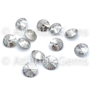 May include: A collection of clear, faceted crystal rhinestones scattered on a white surface. Each rhinestone has a flat, silver backing and a multi-faceted, round top that reflects light. The crystals vary slightly in size and are arranged in a random pattern.