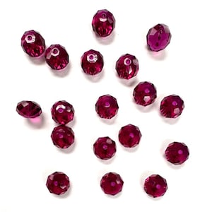 May include: A collection of faceted, deep magenta-colored beads scattered on a white surface. Each bead has multiple facets, creating a sparkling effect. The beads are round with a hole through the center, suggesting they are for jewelry making or crafts.