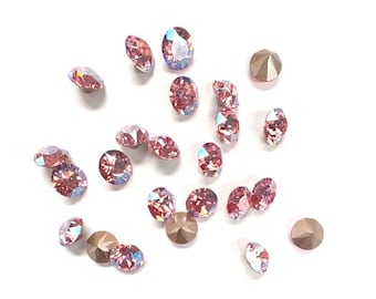 May include: A collection of iridescent pink and gold rhinestones scattered on a white background. The rhinestones are oval and round, with a faceted cut that catches the light. Some are a light gold color.