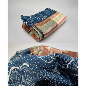 May include: A folded textile featuring a patchwork design with sections of deep blue fabric printed with white floral and fan patterns, alongside panels of muted brown, tan, and green plaid fabric. The material shows visible hand-stitched details.