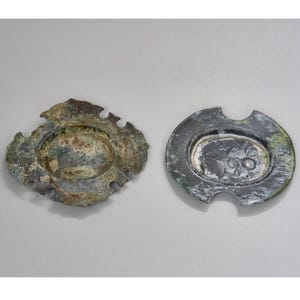 May include: Two antique, weathered metal plates with an oval center and ornate edges. One plate has a scalloped edge, while the other has a rounded shape with cutouts. The plates have a distressed, aged appearance with shades of green and brown.