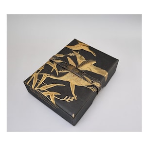 May include: A rectangular black box featuring a gold-colored print of flying cranes and bamboo stalks. The box is secured with a dark metallic strap that includes a central latch mechanism, providing a decorative closure for the storage container.