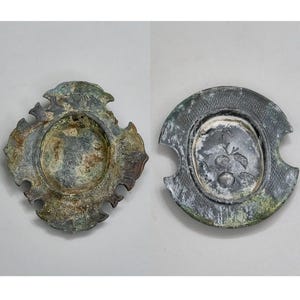 May include: Two antique metal decorative plates with a weathered, aged appearance. One plate has an ornate, floral-like design around the rim. The other plate features a textured rim and a central design of fruit and leaves.