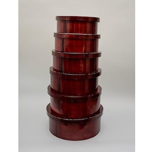 Könnte beinhalten: Ein Stapel von sechs runden, ineinandergreifenden Boxen in verschiedenen Größen. Die Boxen haben eine tiefe, satte burgunderrote Farbe mit glänzendem Finish. Jede Box hat einen Deckel und einen etwas helleren Rand.