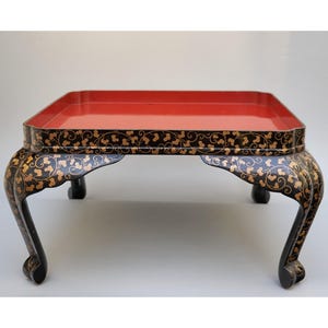 以下が含まれることがあります： 赤色の内側と黒色の外側を持つ正方形のアンティークテーブル。テーブルの外側は金色の花柄で装飾されています。テーブルには4つの湾曲した脚があります。