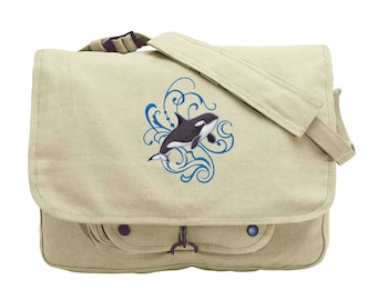 May include: A beige canvas messenger bag with a black and white orca whale design embroidered on the front. The orca is swimming through a blue swirl design. The bag has a flap closure and two pockets on the front.