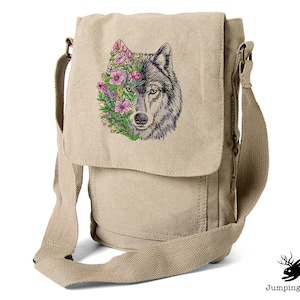 Puede incluir: Una bolsa de mensajero de lona beige con un gráfico de la cara de un lobo. La cara del lobo está rodeada de flores rosas y hojas verdes. La bolsa tiene un bolsillo con cremallera en la parte delantera y una correa larga ajustable.