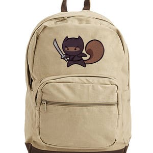 May include: A tan canvas backpack with a brown leather bottom. The backpack has a front pocket with a zipper. The backpack has a black and brown embroidered image of a ninja squirrel holding a sword.