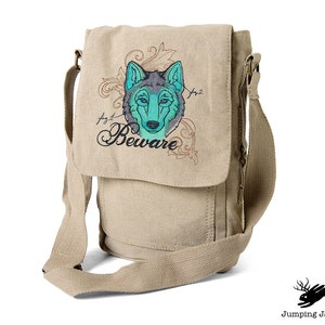 Puede incluir: Bolso bandolera de lona beige con un diseño de cabeza de lobo bordado. El lobo tiene pelo azul y verde y el texto "Beware" está bordado debajo del lobo.
