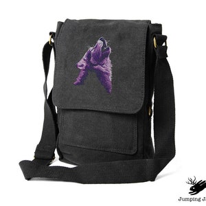 Puede incluir: Una bolsa de mensajero de lona negra con un lobo morado bordado aullando a la luna. La bolsa tiene una correa larga ajustable y una solapa frontal con un bolsillo.