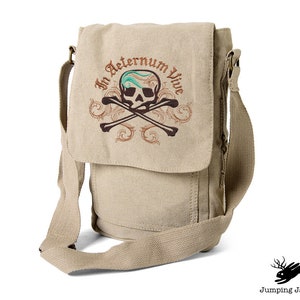 May include: A beige canvas messenger bag with an embroidered skull and crossbones design. The skull has a turquoise bandana and the text "In Aeternum Vive" is embroidered above the skull.