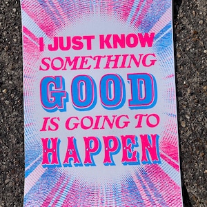 May include: A white poster with pink and blue rays emanating from the centre. The poster has the text "I JUST KNOW SOMETHING GOOD IS GOING TO HAPPEN" in pink and blue letters.
