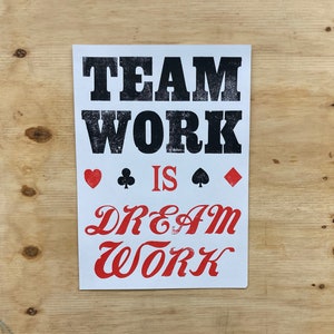 May include: White card with black text that reads "Teamwork is Dream Work". The text is in a bold, blocky font. There are four playing card symbols in red and black below the text.