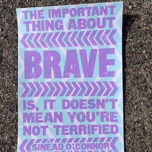 May include: A light blue and purple poster with a quote by Sinead O'Connor. The quote reads "The important thing about brave is, it doesn't mean you're not terrified."