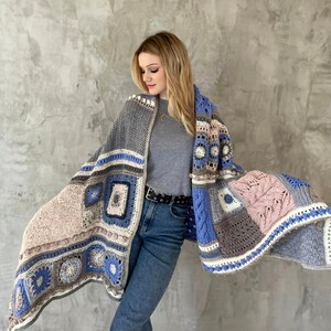 May include: A crocheted shawl with a patchwork design in shades of blue, gray, and pink. The shawl is draped over the shoulders of a person wearing a gray shirt and blue jeans.