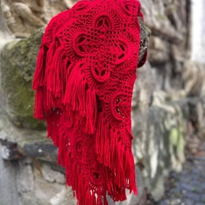 May include: A red crocheted shawl with fringe. The shawl has a delicate pattern and is draped over a stone wall.