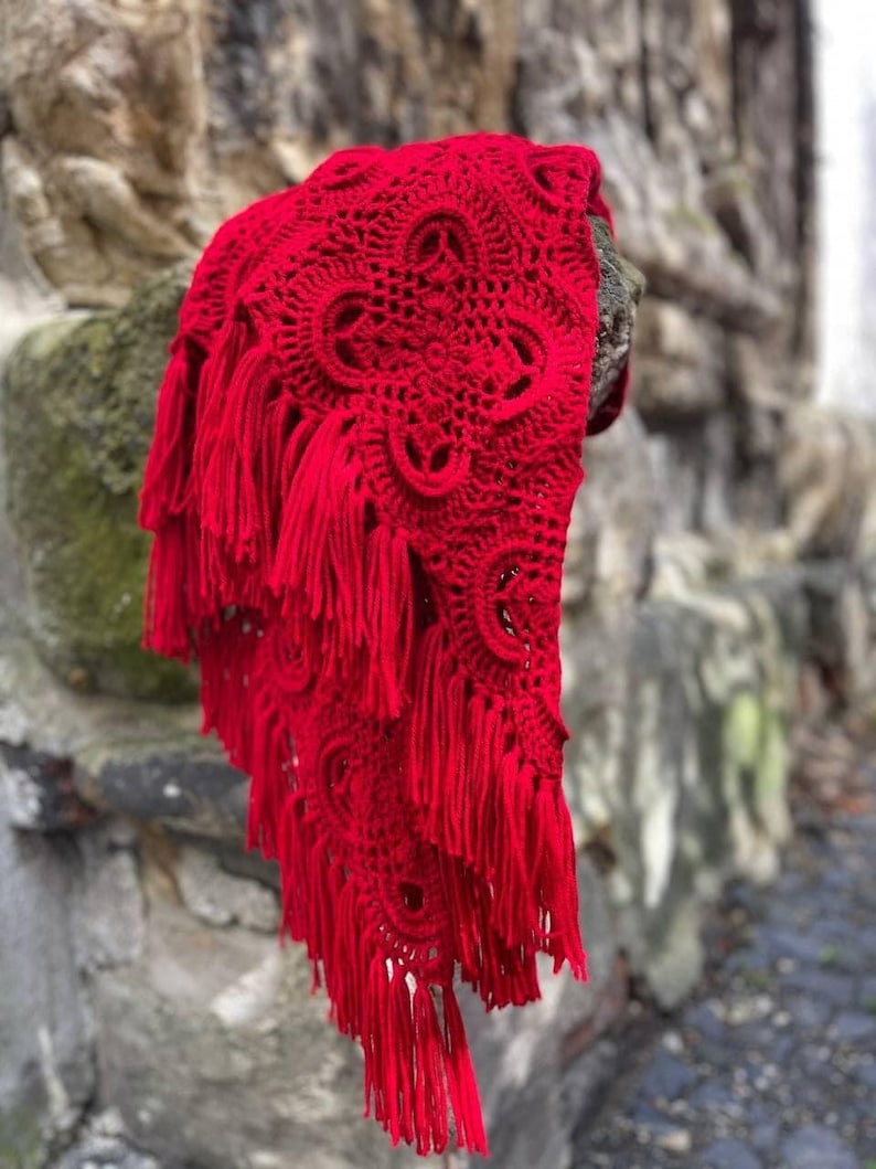 May include: A red crocheted shawl with fringe. The shawl has a delicate pattern and is draped over a stone wall.
