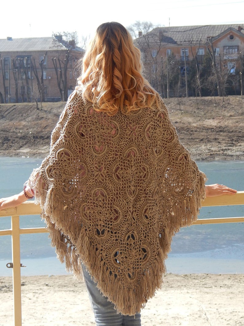 May include: A brown crocheted poncho with fringe. The poncho has a lacy pattern and is worn by a person standing on a balcony overlooking a body of water.