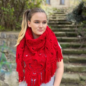 May include: A red crocheted shawl with a lacy pattern and fringe. The shawl is draped around a person's shoulders.