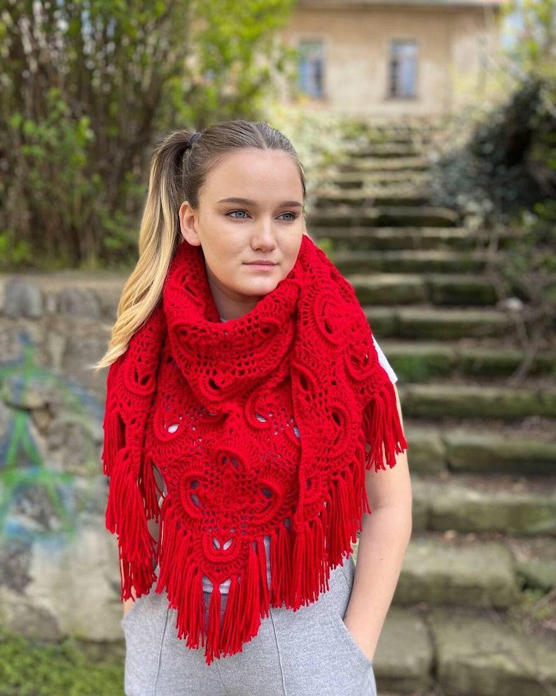 May include: A red crocheted shawl with a lacy pattern and fringe. The shawl is draped around a person's shoulders.