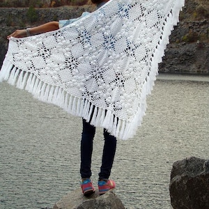 Könnte beinhalten: Ein weißer Häkelschal mit floralem Muster und Fransen. Der Schal wird hochgehalten, um sein detailliertes Muster zu zeigen. Das Bild wurde im Freien aufgenommen, mit Wasser im Hintergrund.