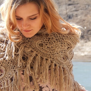 May include: A woman with long blonde hair wearing a brown crocheted shawl with fringe. The shawl is worn over a floral print shirt.