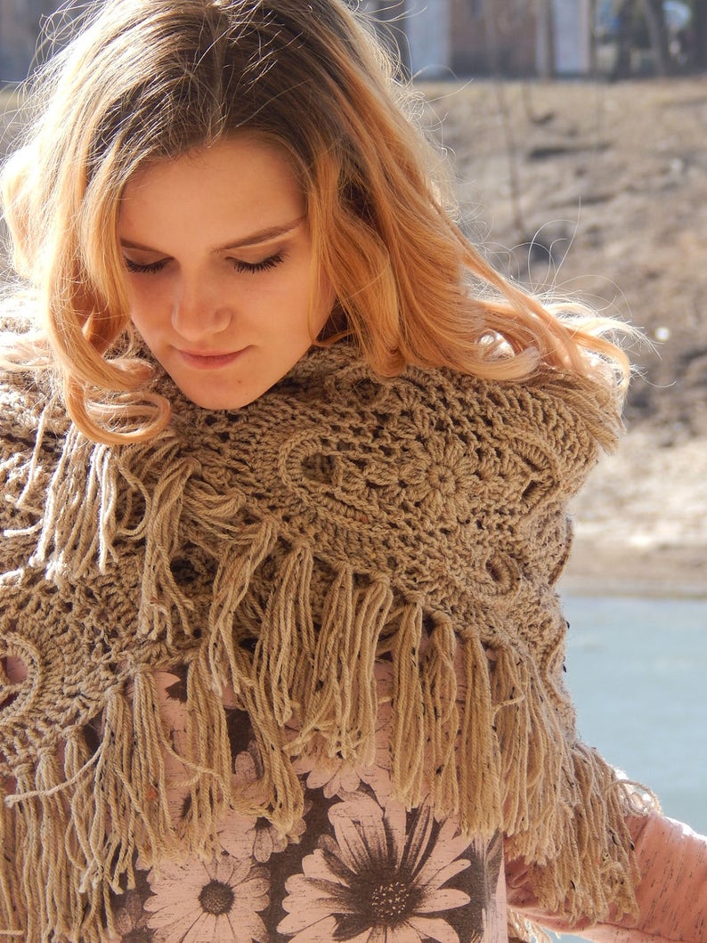 May include: A woman with long blonde hair wearing a brown crocheted shawl with fringe. The shawl is worn over a floral print shirt.