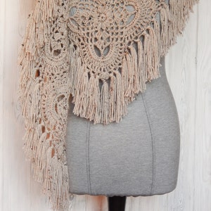 May include: A beige crocheted shawl with a delicate lace pattern and fringe trim. The shawl is draped over a gray dress form.