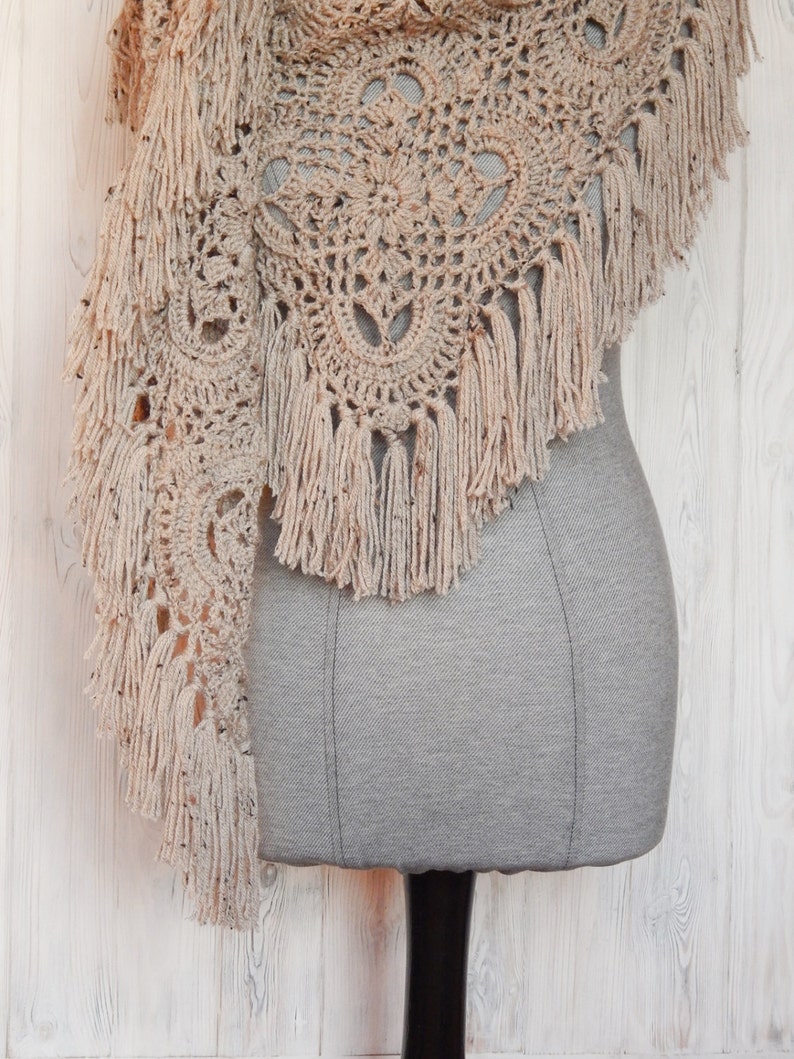 May include: A beige crocheted shawl with a delicate lace pattern and fringe trim. The shawl is draped over a gray dress form.