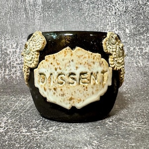 May include: A dark brown ceramic mug featuring a central cream-colored plaque with the word DISSENT embossed in capital letters. The plaque has a speckled texture, and two ornate, textured cream-colored decorative elements flank the sides.