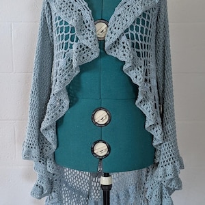 May include: A light blue, open-weave crochet cardigan with a hood and ruffled edges. The garment is displayed on a teal dress form against a white brick wall. The cardigan's design features a delicate, lacy pattern.