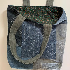 May include: A patchwork denim tote bag with a floral patterned interior. The bag features various shades of blue denim, with white stitching details and gray straps. The interior lining is a dark fabric with small orange and yellow flowers.