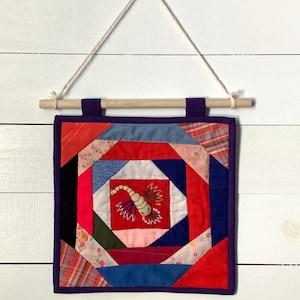 May include: A handmade, square quilt wall hanging with a central red square featuring a scorpion design. The quilt is made of various fabric scraps in red, blue, pink, and patterned fabrics, framed with a purple border and hung with a wooden dowel and string.