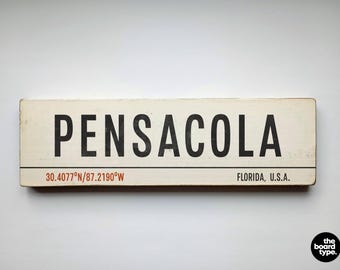 May include: A rectangular off-white wooden sign featuring the black text PENSACOLA in a bold sans-serif font. Below a thin black horizontal line, the text 30.4077°N/87.2190°W appears in orange, with FLORIDA, U.S.A. printed in black on the right side.
