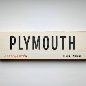 May include: A white wooden sign with black text that reads "PLYMOUTH" and the coordinates "50.3755°N/4.1427°W". Below the coordinates, it reads "DEVON, ENGLAND".