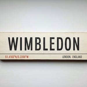 May include: A white wooden sign with black text that reads "WIMBLEDON" and the coordinates "51.4183°N/0.2206°W". Below the coordinates, it reads "LONDON, ENGLAND".