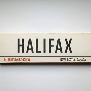 May include: A rectangular off-white wooden sign featuring the black text HALIFAX in a bold sans-serif font. Below a thin horizontal black line, the text 44.8857°N/63.1005°W appears in red, with NOVA SCOTIA, CANADA printed in black to the right.