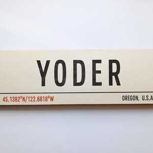 May include: A rectangular wooden sign with the name "YODER" in bold black letters. Below, coordinates and "OREGON, U.S.A." are printed in a smaller font. The sign has a cream-coloured background.