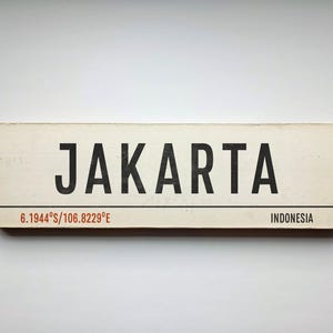Peut inclure: Un panneau rectangulaire en bois blanc cassé avec le texte noir JAKARTA en police sans-serif grasse. Sous une ligne noire horizontale, le texte 6.1944°S/106.8229°E apparaît en rouge, avec le mot INDONESIA imprimé en noir sur la droite.