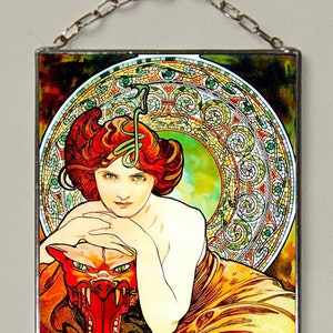 Könnte beinhalten: Ein Kunstwerk aus Buntglas, das eine Frau mit roten Haaren zeigt, die ein rotes, stilisiertes Tier mit Zähnen hält. Die Frau trägt ein gelbes und orangefarbenes Kleid und ist von grünem Laub umgeben. Das Kunstwerk ist mit einer silbernen Kette gerahmt und trägt die Unterschrift des Künstlers "Mucha" in der rechten unteren Ecke.