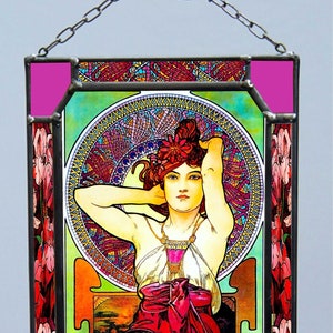 Könnte beinhalten: Ein Buntglasfenster, das eine Frau in einem fließenden Kleid mit einem floralen Design zeigt. Die Frau ist von rosa und roten Iris umgeben. Das Fenster ist in Schwarz mit rosa Rändern gerahmt.