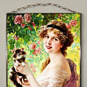 May include: A rectangular glass wall hanging featuring a printed illustration of a person with curly brown hair and a black headband holding a fluffy brown and white kitten. The background shows pink roses and green foliage with a metal chain at the top.